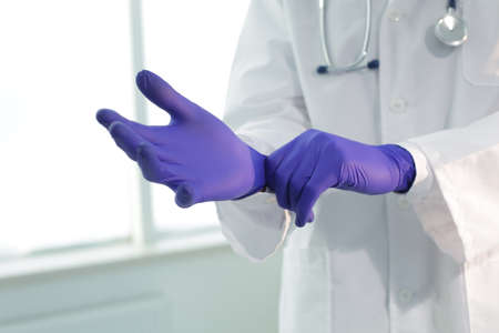 Doctor with stethoscope in a medical gown puts on protective gloves in a clinicの写真素材