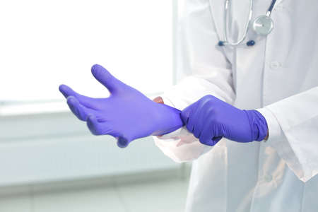 Doctor with stethoscope in a medical gown puts on protective gloves in a clinicの写真素材