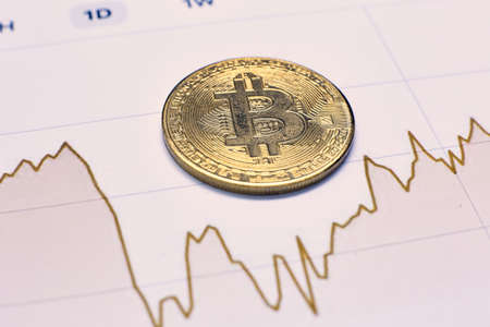 Gold coin of bitcoin cryptocurrency and charts of changes in cryptocurrency rates lie on a wooden table in the officeの写真素材