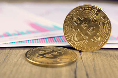 One gold coin of the bitcoin cryptocurrency stands on its own end, the second one lies on the charts...
