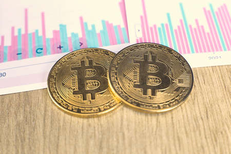 Two gold coins of bitcoin cryptocurrency and charts of changes in cryptocurrency rates lie on a wooden table in the officeの写真素材