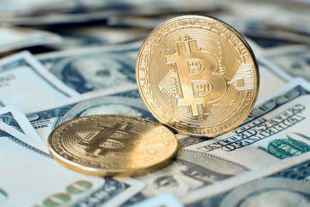 The gold bitcoin cryptocurrency coin stands on its own end, the second one lies on one hundred dollar billsの写真素材