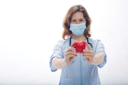 Doctor woman stands in a protective mask with a stethoscope and holds a heart in her hands isolated...