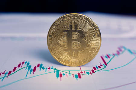 The gold bitcoin cryptocurrency coin stands on its own end. The graph of changes in cryptocurrency rates lies on a wooden table in the officeの写真素材