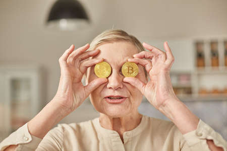 An elderly woman holds bitcoin coins in her hands near her eyes. Senior citizen happy life conceptの写真素材