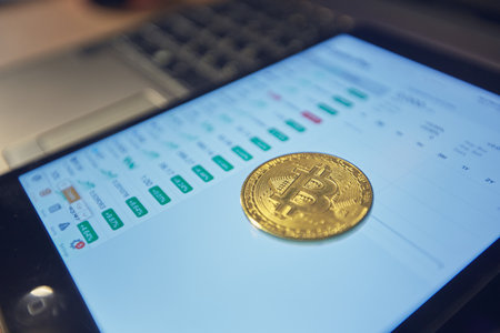 A man holds a bitcoin coin in his hand. The graphs of changes in cryptocurrency rates are shown on the laptop monitorの写真素材