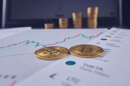 Two bitcoins are on the graphs of cryptocurrency rate changes on a laptop. In the background, several rows of coins and a laptop screen.の写真素材
