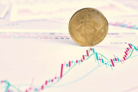 The gold bitcoin cryptocurrency coin stands on its own end. The graph of changes in cryptocurrency rates lies on a wooden table in the officeの写真素材