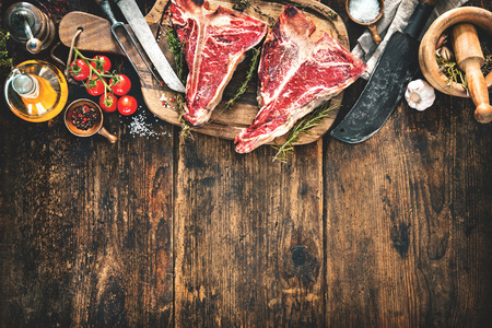 Raw dry aged t-bone steaks for grill with fresh herbs, vegetables and cleaver on rustic wooden boardの写真素材