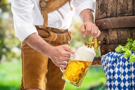 Bavarian man in leather trousers is pouring a large lager beer in tap from wooden beer barrel in the garden. Background for Oktoberfest, folk or beer festivalの写真素材
