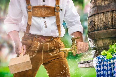 Bavarian man in leather trousers taps a wooden barrel of beer in the garden. Background for Oktoberfest, folk or beer festivalの写真素材