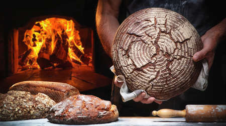 Baker's hands holding and presenting fresh baked loaf of bread in front of a burning oven
