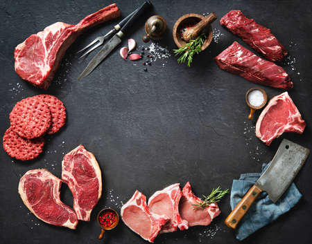 Variety of raw cuts of meat, dry aged beef steaks and hamburger patties for grilling with seasoning and utensils on dark rustic boardの写真素材