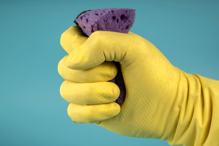 Hand in yellow rubber glove holds sponge for washing dishes and cleaning, hand clenches sponge into fist on blue backgroundの写真素材