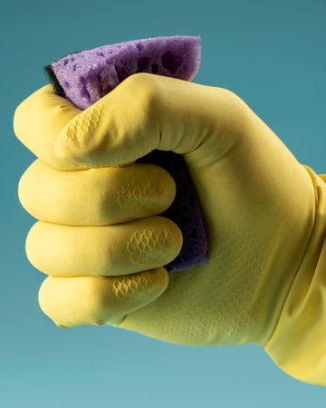 Hand in yellow rubber glove holds sponge for washing dishes and cleaning, hand clenches sponge into...