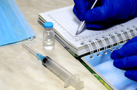 There is a syringe and a medical mask on the table, and the vaccine is on the table. The doctor makes notes in a notebook, his hands are protected by blue gloves. Concept - pandemic, covid, vaccination.の写真素材