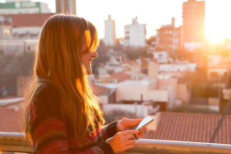 Woman with a phone in her hands. She's at the sunset with a smartphone. The city at the background. Retro style colors. Woman in Europe.の写真素材