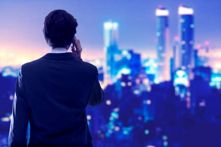 Young businessman on his back and talking on the mobile phone while looking the city at night.の写真素材