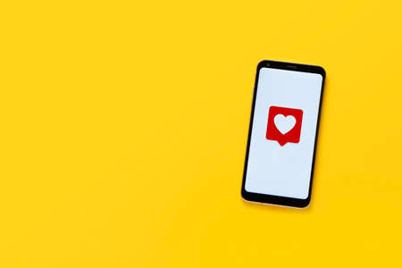 Top view of cell phone with heart emoticon. Minimalist design. White screen and yellow background, copy space. Social network concept.の写真素材