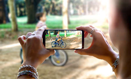Woman photographing her little son with a cell phone. The child is learning to ride a bicycle on the...
