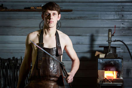 portrait of a young blacksmith in a leather uniform works in a smithyの写真素材