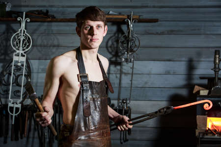 portrait of a young blacksmith in a leather uniform works in a smithyの写真素材