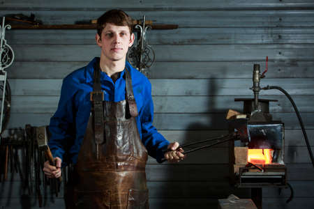 portrait of a young blacksmith in a leather uniform works in a smithyの写真素材