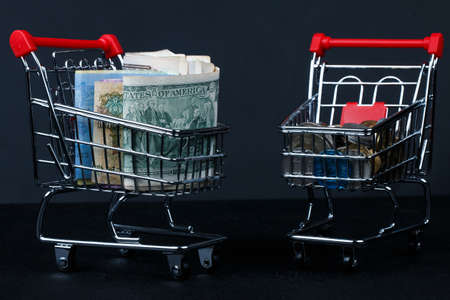 Selective focus on  coins in mini shopping cart on pile of  banknote on dark black background as shopping concept.の写真素材
