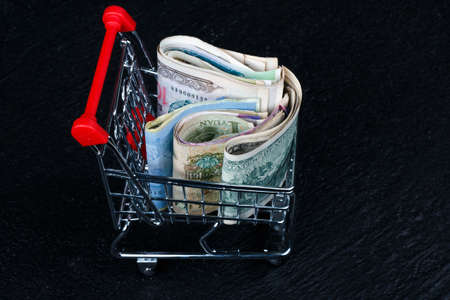 Selective focus on  coins in mini shopping cart on pile of  banknote on dark black background as shopping concept.の写真素材
