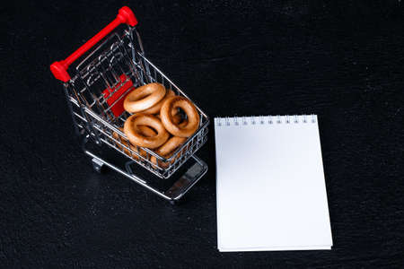 white notebook (mock up design) workup space, shopping list  shopping cart, bagel, corn on dark...
