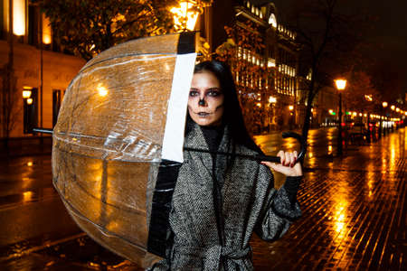 beautiful girl with long black hair is holding a transparent umbrella. It's evening in the street,raining. She wears a gray coat. On the face a make-up with the image of a skeleton.Gold backgroundの写真素材