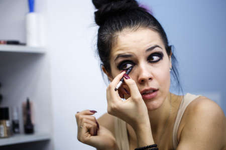 Beautiful  Woman with dark hair Looking Into The Mirror At Herself And Applying black shadowsの写真素材