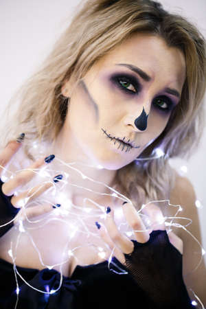 portrait of a beautiful young blond girl with bright make-up in the style of Halloween. She holds burning garlands and looks at the cameraの写真素材