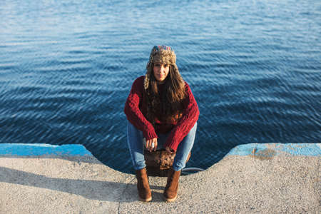 Young woman in hat and casual clothes sitting on pier and looking at cameraの写真素材