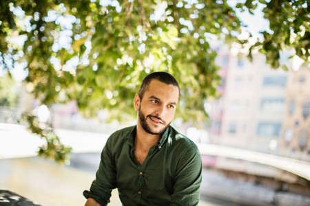 Young bearded man sitting outdoors under trees and smiling while looking away