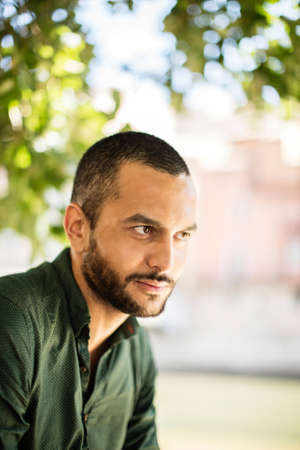 Young bearded man sitting outdoors under trees and smiling while looking away