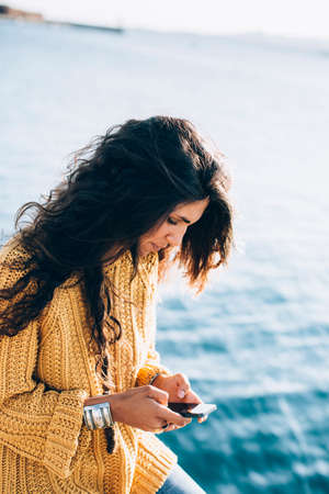 Smiling young woman sitting on pier and chatting over phone. Summertimeの写真素材