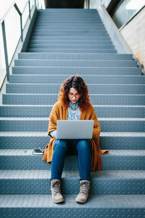 Young red haired woman sitting on stairs while using laptopの写真素材