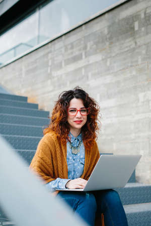 Young red haired woman sitting on stairs while using laptop looking at camera