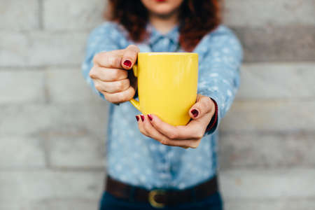 Close-up of woman's hands holding yellow cup. Unfocused background on cement wall. Unrecognizableの写真素材