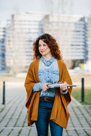 Smiling curly haired woman walking the street while holding laptop. Cityscape on background