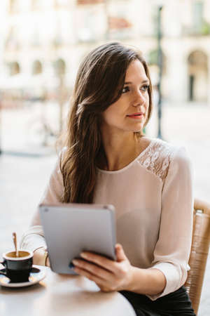 Pretty young female in formal outfit smiling and using tablet while sitting at cafe.の写真素材