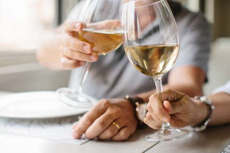 Crop faceless shot of mature couple clinking wineglasses with white wine in restaurant.の写真素材