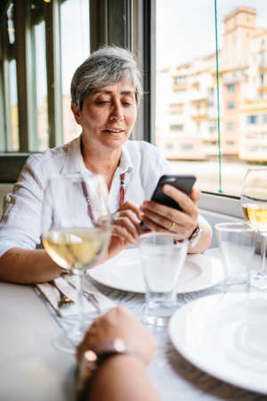 Beautiful senior lady in casual clothing browsing smart phone while sitting in restaurant.の写真素材