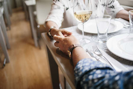 Crop faceless shot of mature couple holding hands while sitting at table in restaurant.の写真素材