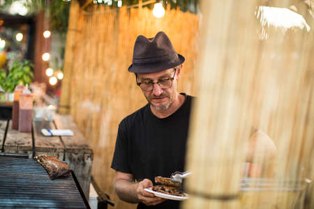 Man in glasses and hat roasting meat on grill outside.の写真素材