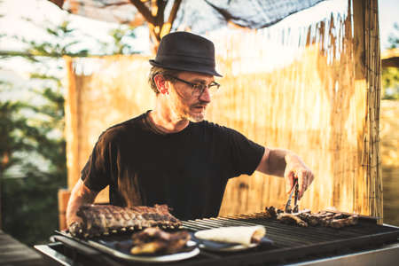 Man in glasses and hat roasting meat on grill outside.の写真素材