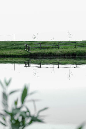Calm rural pond with a wooden fence along the bank and reflections under a bright sky.の写真素材