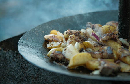 Savory outdoor-cooked meal of fried potatoes, bacon, and onions, sizzling on a black metal disc.の写真素材