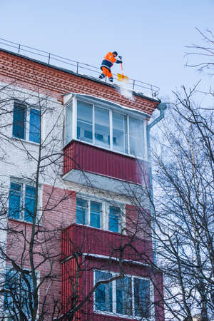 Parkovaya Street, Balashikha, Russia-February 19, 2021. Snow removal. A worker removes snow with a shovel from the roof of a building after a snowfall.のeditorial素材